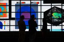 Three people stand in front of a large screen displaying various scientific and mathematical visualizations. The screen shows multiple graphs, charts, and images with color-coded data, including heat maps and 3D models. The figures are silhouetted against the brightly lit display.