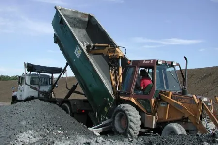 A construction scene showing a backhoe loader with a front-end loader attachment lifting the bed of a green dump truck, which is unloading gravel or dirt onto the ground. Another white dump truck is parked nearby, partially visible in the background. The sky is clear and blue.